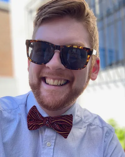 Kyle smiling in sunglasses, a light blue shirt and a patterned red bowtie